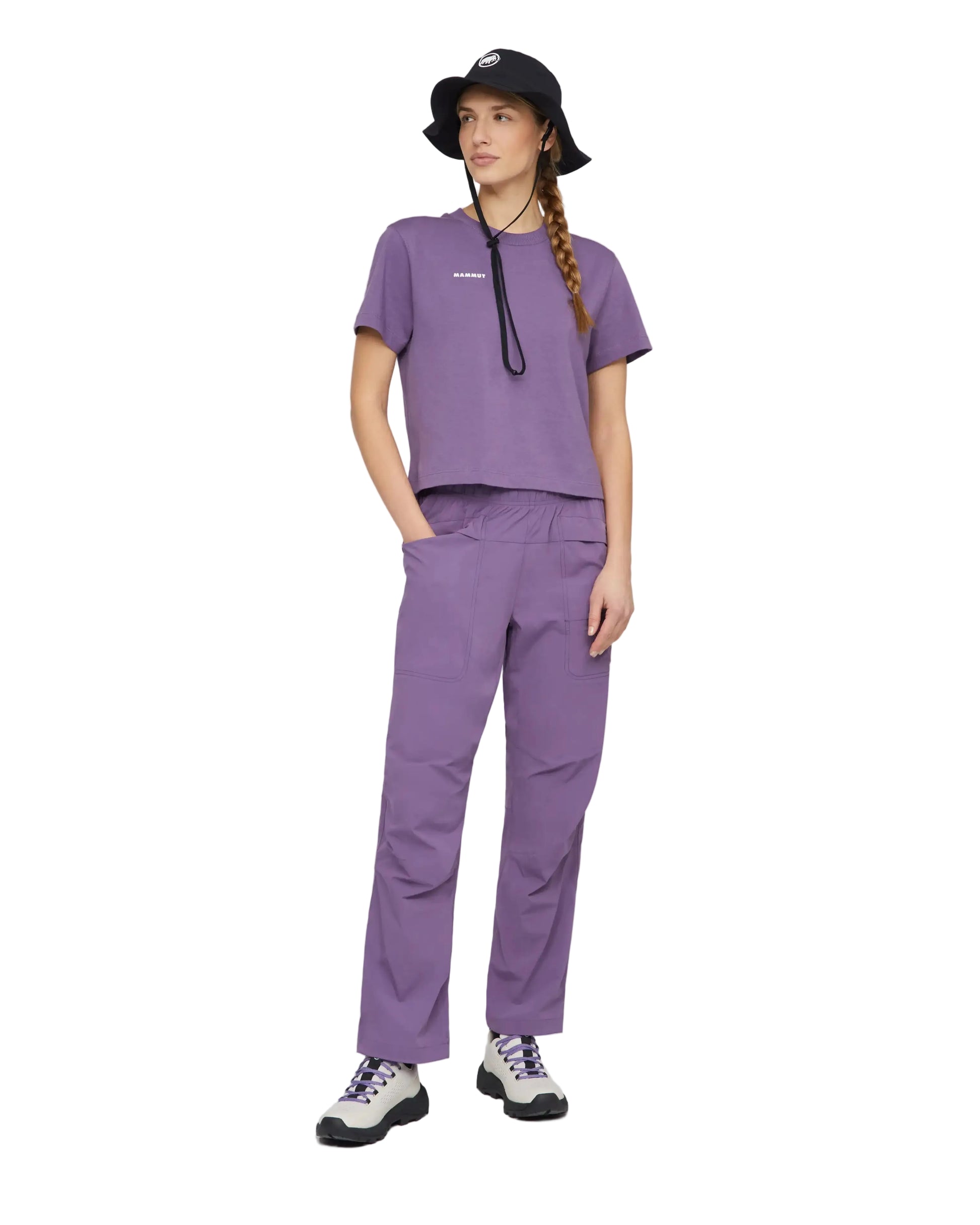 A model wearing a matching purple set consisting of a short-sleeve cropped top and the mammut sender pants, styled with a black bucket hat.